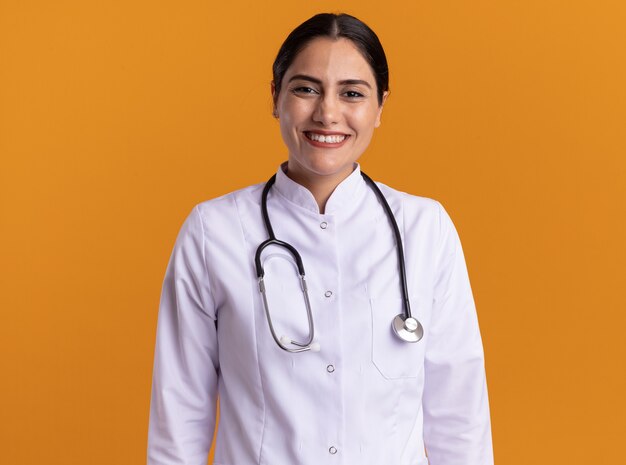 Felice giovane donna medico in cappotto medico con lo stetoscopio intorno al collo guardando la parte anteriore con un grande sorriso sul viso in piedi sopra la parete arancione