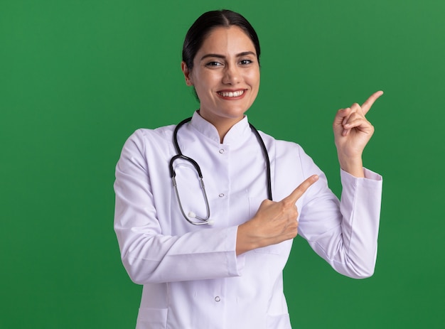 Felice giovane donna medico in cappotto medico con uno stetoscopio guardando davanti con il sorriso sul viso rivolto con le dita indice al lato in piedi sopra la parete verde