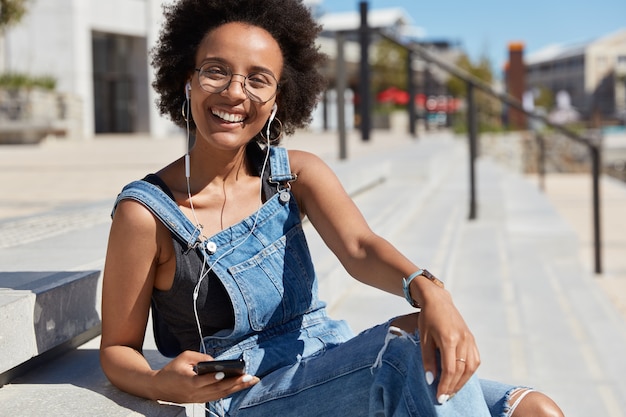 Felice giovane donna nera e rilassata ascolta la musica preferita o la trasmissione radiofonica, ride allegramente, indossa abiti casual, occhiali trasparenti, modelli all'aperto in strada.