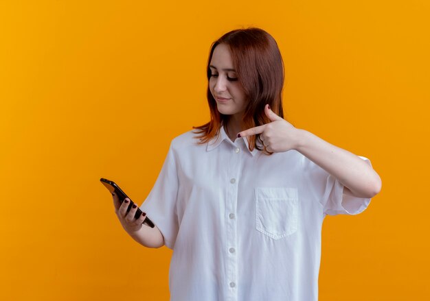 felice giovane donna redhead holding e punti al telefono isolato su giallo