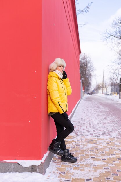 Felice giovane donna su uno sfondo di un muro rosso in vestiti caldi in una giornata di sole invernale sorridente e parlando al telefono su un marciapiede innevato della città