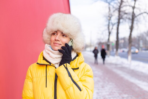 Felice giovane donna su uno sfondo di un muro rosso in vestiti caldi in una giornata di sole invernale sorridente e parlando al telefono su un marciapiede innevato della città