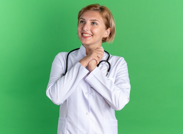 Felice giovane dottoressa che indossa un abito medico con uno stetoscopio che si tiene per mano insieme isolato sul muro verde