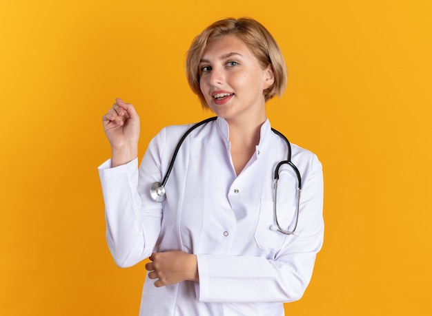 Felice giovane dottoressa che indossa una tunica medica con uno stetoscopio isolato sulla parete arancione