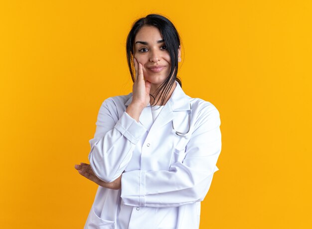 Felice giovane dottoressa che indossa una veste medica con uno stetoscopio che mette la mano sulla guancia isolata sulla parete gialla