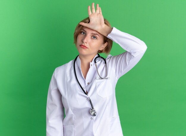 Felice giovane dottoressa che indossa una veste medica con uno stetoscopio che mette la mano sulla testa isolata sulla parete verde