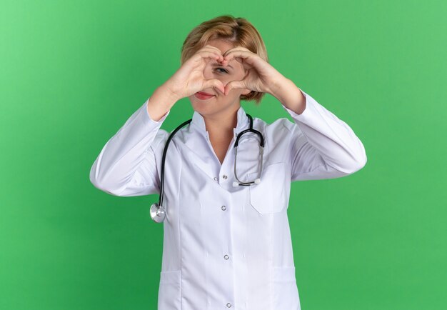 Felice giovane dottoressa che indossa una veste medica con uno stetoscopio che mostra il gesto del cuore isolato sulla parete verde