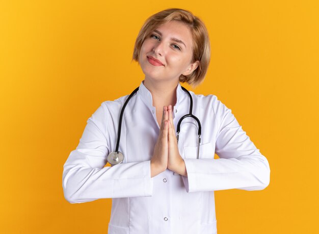 Felice giovane dottoressa che indossa una veste medica con uno stetoscopio che mostra il gesto di preghiera isolato sulla parete arancione