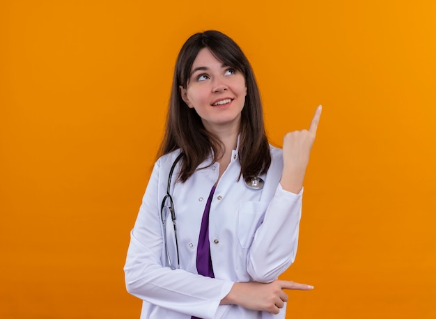 Felice giovane dottoressa in abito medico con lo stetoscopio punta verso l'alto e guarda su sfondo arancione isolato con spazio di copia