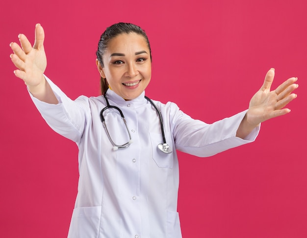 Felice giovane dottoressa in camice bianco con stetoscopio intorno al collo che fa un gesto di benvenuto che apre le mani in piedi sul muro rosa