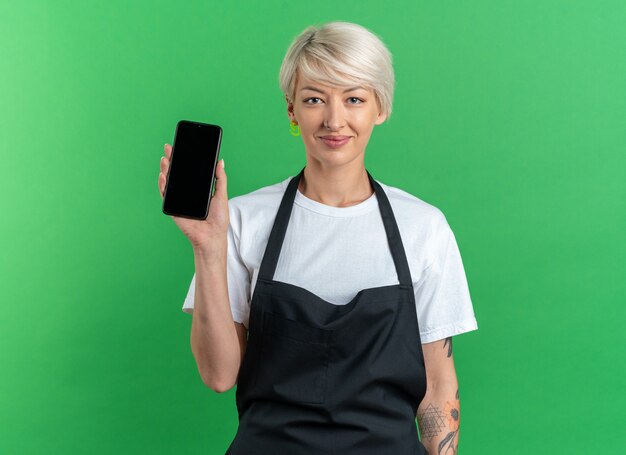 Felice giovane e bella barbiere in uniforme che tiene il telefono isolato sul muro verde