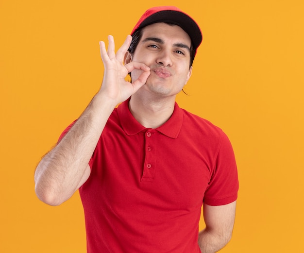 Felice giovane fattorino caucasico in uniforme blu e berretto che tiene la mano dietro la schiena facendo un gesto delizioso isolato sul muro arancione orange