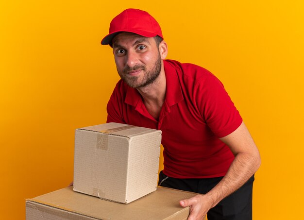 Felice giovane fattorino caucasico in uniforme rossa e berretto piegato tenendo le mani sulla scatola di cartone guardando la telecamera isolata sulla parete arancione