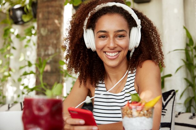 Felice giovane femmina afroamericana positiva legge il messaggio sul cellulare, ascolta l'audio con le cuffie, riposa dopo il lavoro in un accogliente ristorante, gode di connessione internet gratuita. Tempo libero e riposo