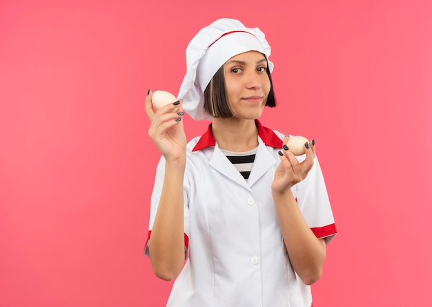 Felice giovane femmina cuoco in uniforme da chef tenendo le uova isolate sulla parete rosa