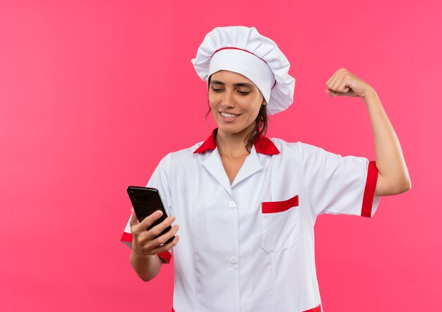Felice giovane femmina cuoco indossando chef uniforme holding e guardando il telefono che mostra un forte gesto sulla parete rosa isolata con lo spazio della copia