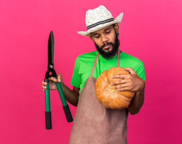 Felice giovane giardiniere afro-americano che indossa un cappello da giardinaggio tenendo la zucca con le forbici