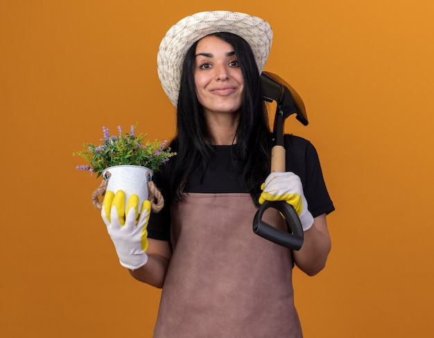 Felice giovane giardiniere caucasica ragazza che indossa uniforme e cappello con guanti da giardiniere che tengono vanga e vaso di fiori isolati su parete arancione