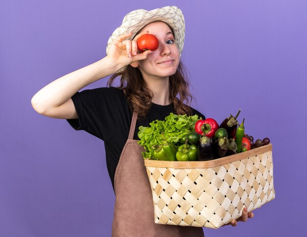 Felice giovane giardiniere femmina che indossa cappello da giardinaggio tenendo cesto di verdure che mostra gesto di sguardo con pomodoro