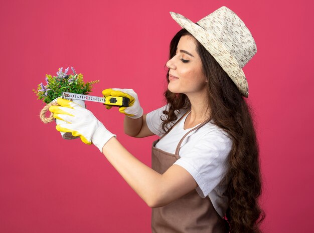 Felice giovane giardiniere femminile in uniforme che indossa guanti e cappello da giardinaggio misurazione vaso di fiori con metro a nastro isolato sulla parete rosa con spazio di copia