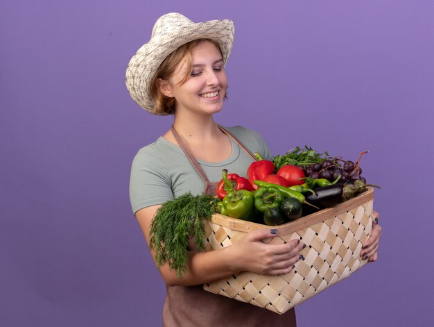 Felice giovane giardiniere femminile slavo che indossa cappello da giardinaggio tenendo e guardando il cesto di verdure sulla porpora