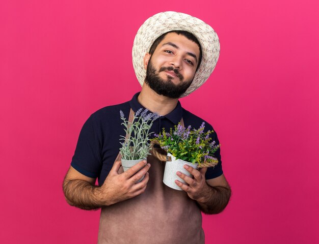 Felice giovane giardiniere maschio caucasico che indossa il cappello da giardinaggio che tiene i vasi da fiori isolati sulla parete rosa con lo spazio della copia