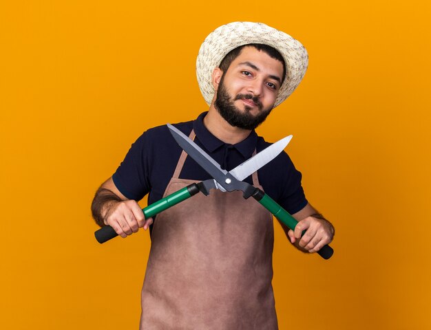 Felice giovane giardiniere maschio caucasico indossando il cappello da giardinaggio tenendo le forbici da giardinaggio isolate sulla parete arancione con lo spazio della copia