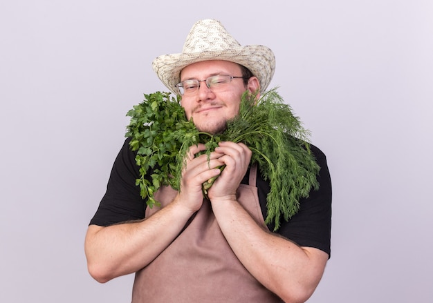 Felice giovane giardiniere maschio che indossa cappello da giardinaggio che tiene aneto con coriandolo intorno al viso isolato sul muro bianco