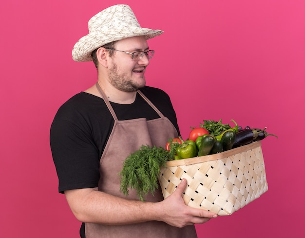 Felice giovane giardiniere maschio che indossa cappello da giardinaggio tenendo e guardando il cesto di verdure isolato sulla parete rosa