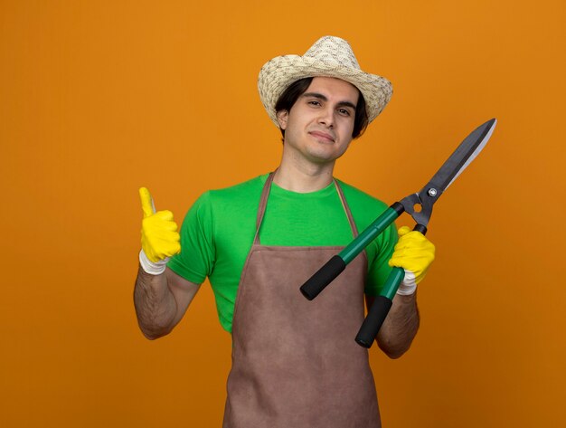 Felice giovane giardiniere maschio in uniforme che indossa il cappello da giardinaggio con i guanti che tengono i clippers che mostrano il pollice in su isolato sull'arancio