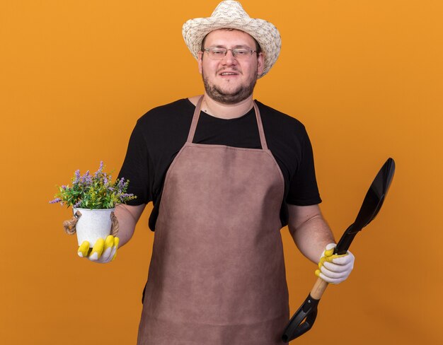 Felice giovane giardiniere maschio indossando guanti e cappello da giardinaggio tenendo la vanga con il fiore in vaso di fiori isolato sulla parete arancione