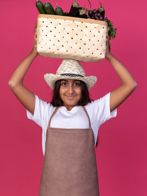 Felice giovane giardiniere ragazza in grembiule e cappello estivo azienda cassa piena di verdure sopra la sua testa con il sorriso sul viso in piedi sopra il muro rosa