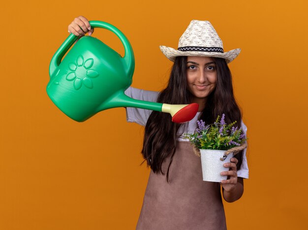 Felice giovane giardiniere ragazza in grembiule e cappello estivo che tiene annaffiatoio e pianta in vaso di irrigazione con il sorriso sul viso in piedi sopra la parete arancione