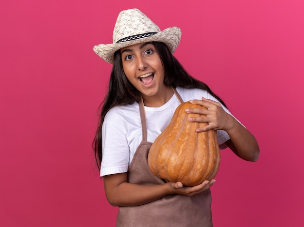 Felice giovane giardiniere ragazza in grembiule e cappello estivo tenendo la zucca sorridente allegramente in piedi sopra la parete rosa