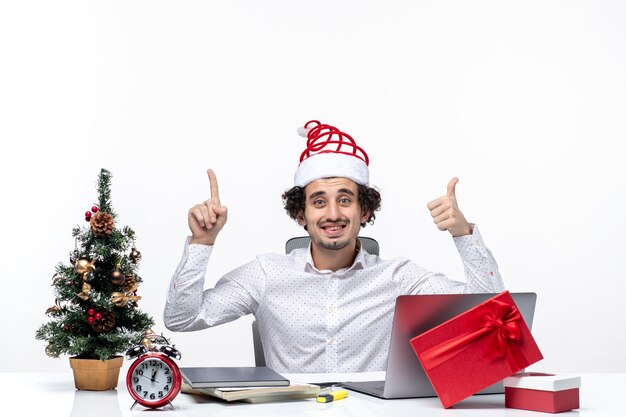 Felice giovane imprenditore con buffo cappello di Babbo Natale che punta sopra e che fa il gesto giusto in ufficio su sfondo bianco