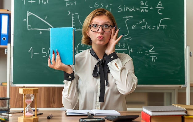 Felice giovane insegnante con gli occhiali si siede a tavola con gli strumenti della scuola che tiene il libro in classe