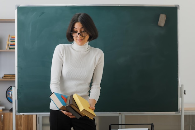 felice giovane insegnante femminile che indossa occhiali tenendo e guardando i libri in piedi davanti alla lavagna in classe