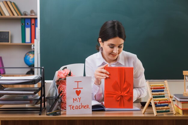 felice giovane insegnante femminile che tiene e guarda il regalo seduto alla scrivania con gli strumenti della scuola in classe