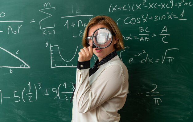 Felice giovane insegnante femminile con gli occhiali in piedi davanti alla lavagna guardando la telecamera con la lente di ingrandimento in aula