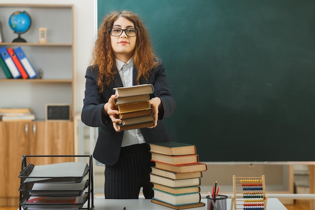 felice giovane insegnante femminile in piedi davanti alla lavagna che porge libri alla telecamera in classe