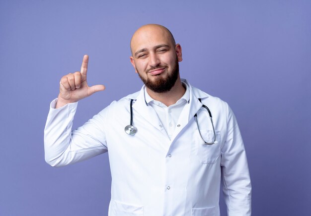 Felice giovane maschio calvo medico indossando abito medico e stetoscopio fingendo di tenere qualcosa di isolato su sfondo blu