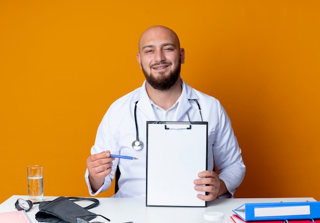 Felice giovane maschio calvo medico indossando abito medico e stetoscopio seduto alla scrivania con strumenti medici holding e punti con penna negli appunti isolato su sfondo arancione