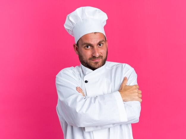 Felice giovane maschio caucasico cuoco in uniforme da chef e berretto in piedi con postura chiusa guardando la telecamera isolata sulla parete rosa