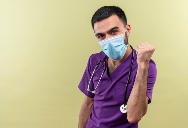 Felice giovane maschio medico indossando viola chirurgo abbigliamento e stetoscopio mascherina medica che mostra sì gesto su sfondo verde isolato