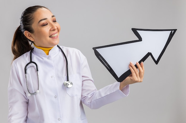 Felice giovane medico donna in camice bianco medico con stetoscopio intorno al collo tenendo la freccia guardandolo sorridendo allegramente