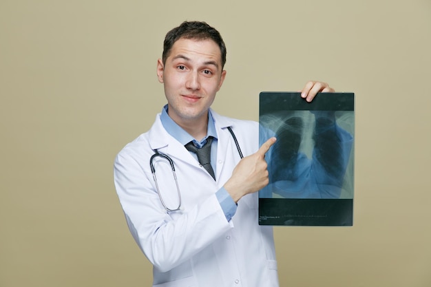 Felice giovane medico maschio che indossa accappatoio medico e stetoscopio intorno al collo guardando la fotocamera che mostra xray shot puntando il dito su di esso isolato su sfondo verde oliva