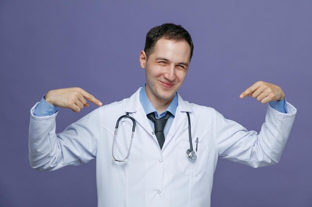 Felice giovane medico maschio che indossa un abito medico e uno stetoscopio intorno al collo guardando la fotocamera che punta a se stesso isolato su sfondo viola