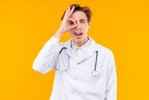 Felice giovane medico maschio che indossa una tunica medica con uno stetoscopio che mostra il gesto di sguardo isolato sulla parete arancione