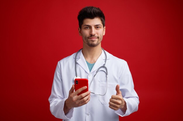 Felice giovane medico maschio che indossa uniforme medica e stetoscopio intorno al collo tenendo il telefono cellulare guardando la fotocamera che mostra il pollice in su isolato su sfondo rosso