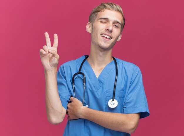 Felice giovane medico maschio sbattuto le palpebre che indossa l'uniforme del medico con lo stetoscopio che mostra il gesto di pace isolato sulla parete rosa
